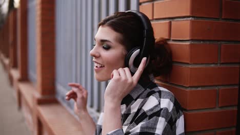 hermosa morena apoyada en una valla de ladrillo al aire libre, escuchando música en auriculares con los ojos cerrados. siente la música. gestando emocionalmente