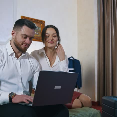 Smiling couple looking into laptop. Young beautiful woman and handsome guy sitting on a bed in hotel room and using laptop together. Romantic couple in hotel