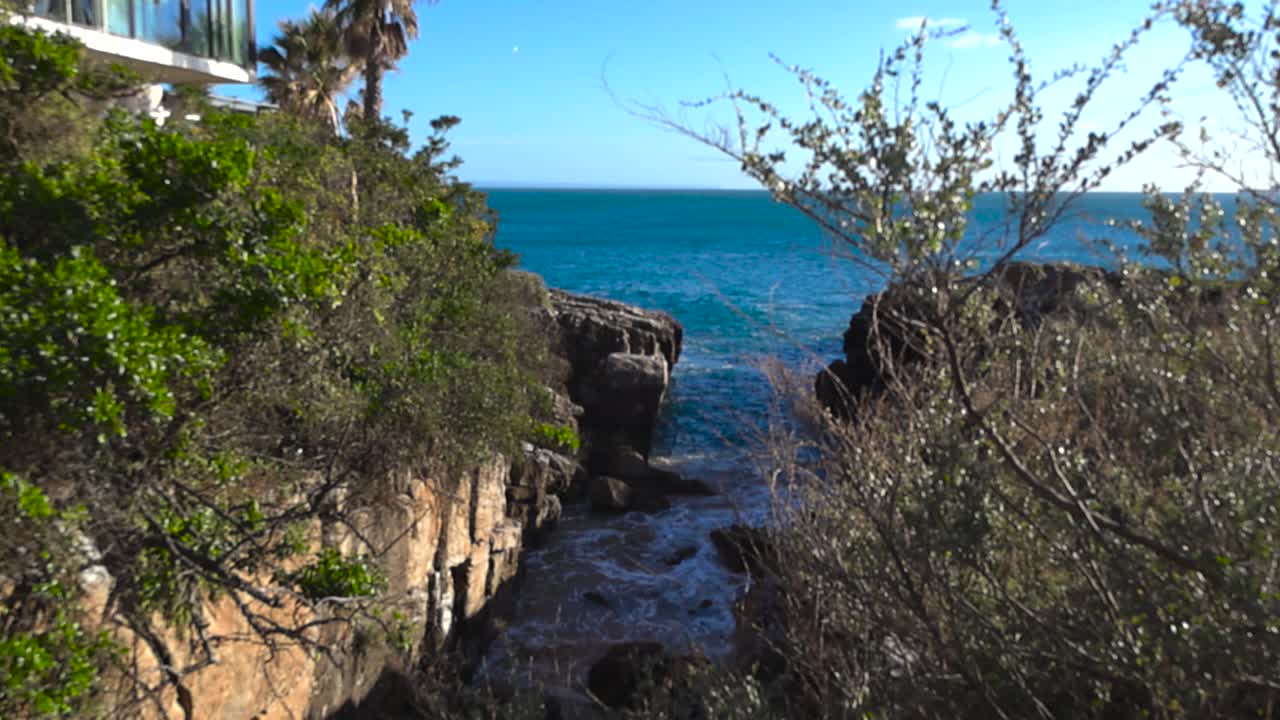 Scenario costiero mozzafiato con acqua oceanica turchese che si riversa in una gola rocciosa in una luminosa giornata di sole. Le aspre scogliere sono coperte da una vegetazione verde lussureggiante sotto un cielo azzurro e limpido.