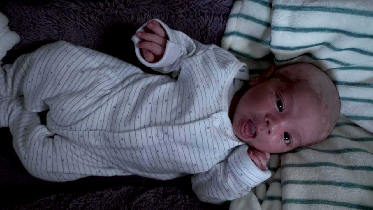Neonato sdraiato nel letto che indossa un pigiama intero bianco, mentre si muove con la bocca aperta, con un taglio di capelli recente.