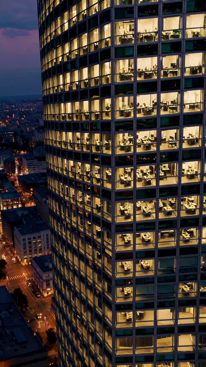 Aerial video view of a lit office building at dusk, showcasing a bustling city life