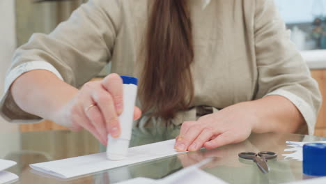 Woman applying glue between paper craft on table with scissors and other supplies visible in background, creating DIY project