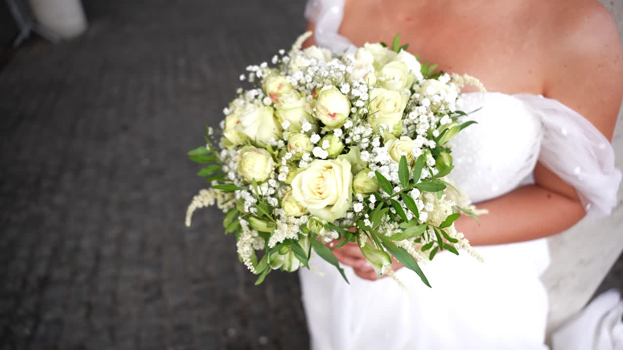 Sposa in abito bianco che tiene un elegante bouquet di rose bianche, gypsophila e verde, perfetto per catturare dettagli romantici di matrimonio