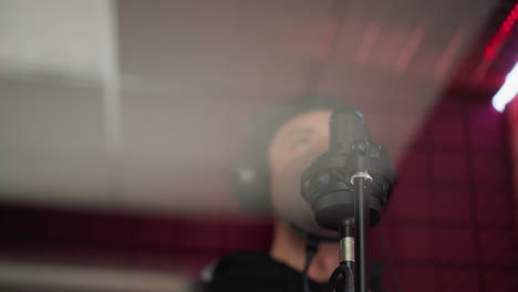 blurry shot of a man dressed in black, wearing headphones and standing in front of a microphone in a red soundproofed room. focuses on the microphone, with the singer in the background