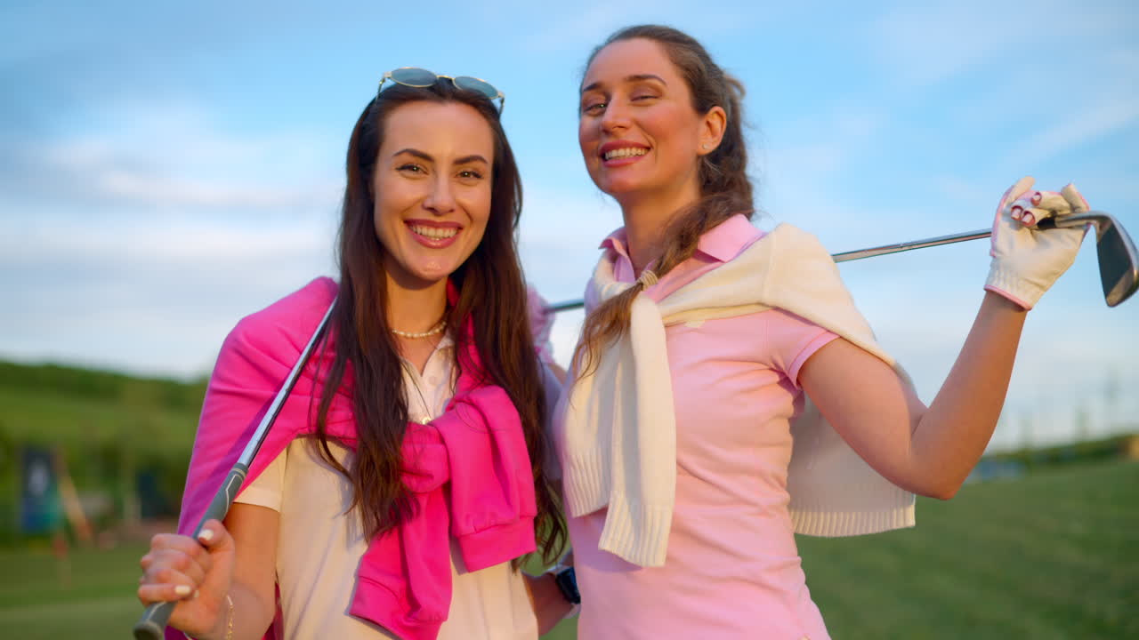 Two women in white and pink clothes, posing with golf clubs in their hands, on a grass field