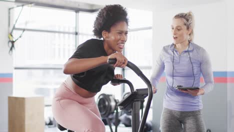 video of diverse female fitness trainer and woman on exercise bike working out at a gym