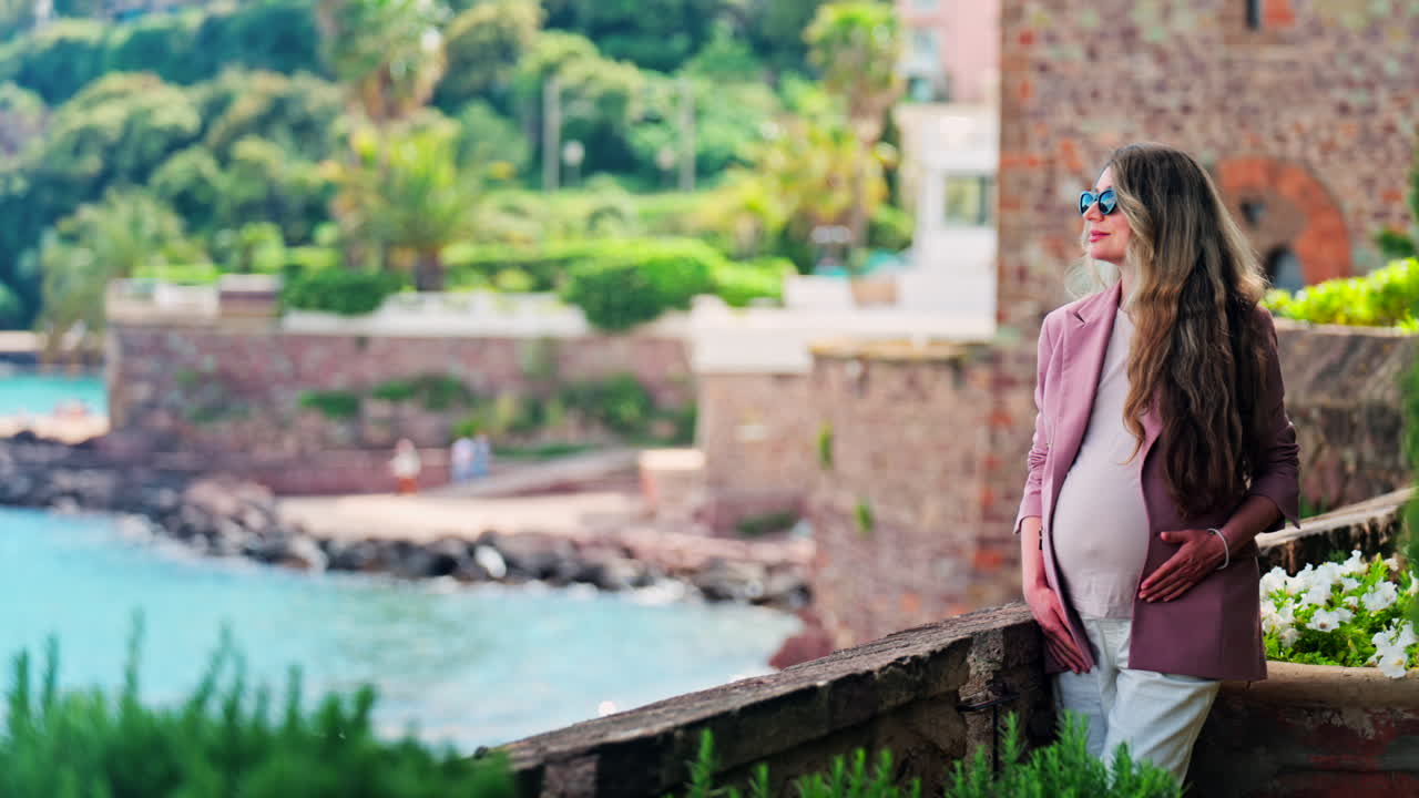 Pregnant woman with long hair, wearing sunglasses standing with the Chateau de la Napoule Castle in Mandelieu-La Napoule, France in the background