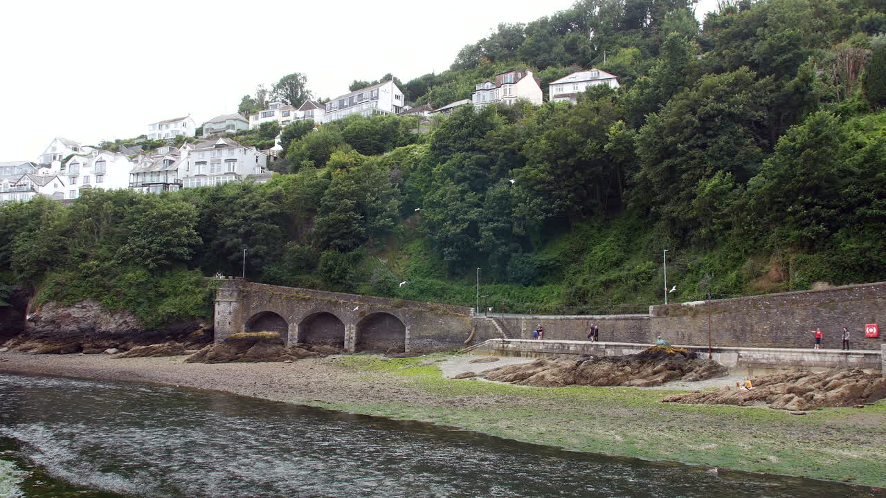 An arched wall runs along a rocky shoreline beneath a steep, tree-covered hillside, with houses built above and a few people walking near the waterfront in Looe, Cornwall, England