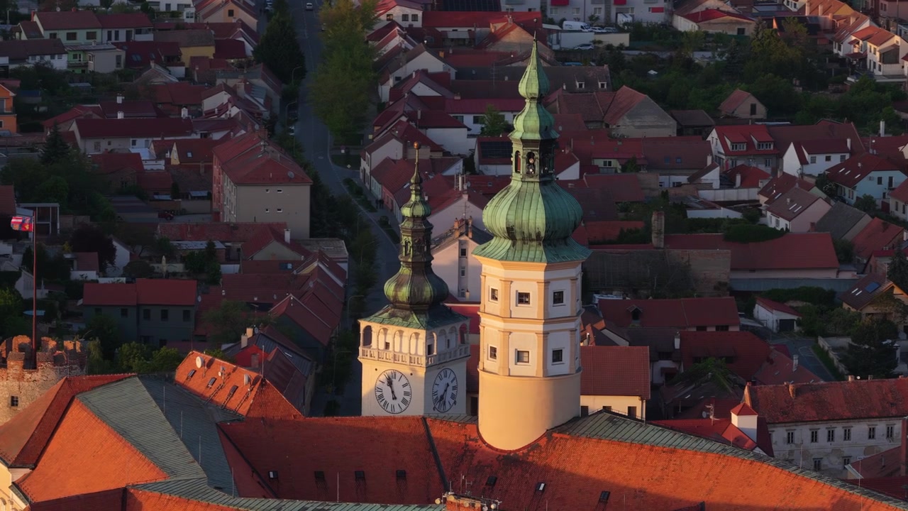 Vista aerea con drone di due torri di chiesa barocche con cupole a cipolla in rame verde e quadranti dell'orologio che brillano nella calda luce del tramonto serale sopra tetti di tegole rosse, Mikulov, Moravia, Repubblica Ceca