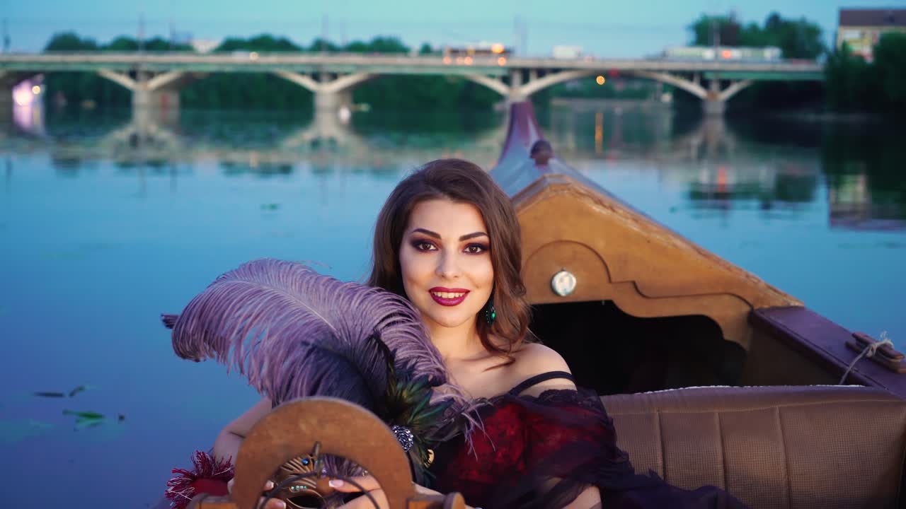A charming woman with a bright make-up is holding a violet feather in her hand and smiling in the evening on the gondola. Blurred Background