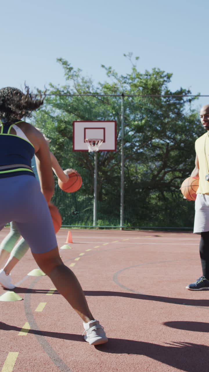 Vertical video of diverse female basketball team training on sunny court, in slow motion