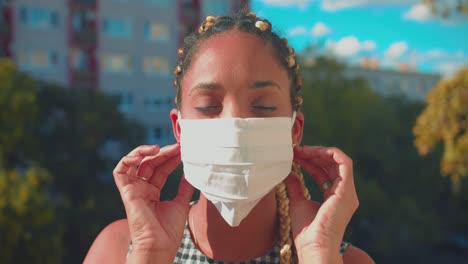 portrait of serious young mixed race woman with hair braids taking off face mask in exterior on sunny summer day, looking at camera, full frame