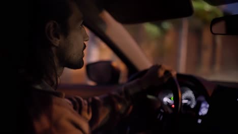 disparado desde el lado un hombre con una barba morena conduce un coche moderno mientras está sentado en la cabina y conduciendo a través de la ciudad de la noche. conducir un coche por la noche y por la noche
