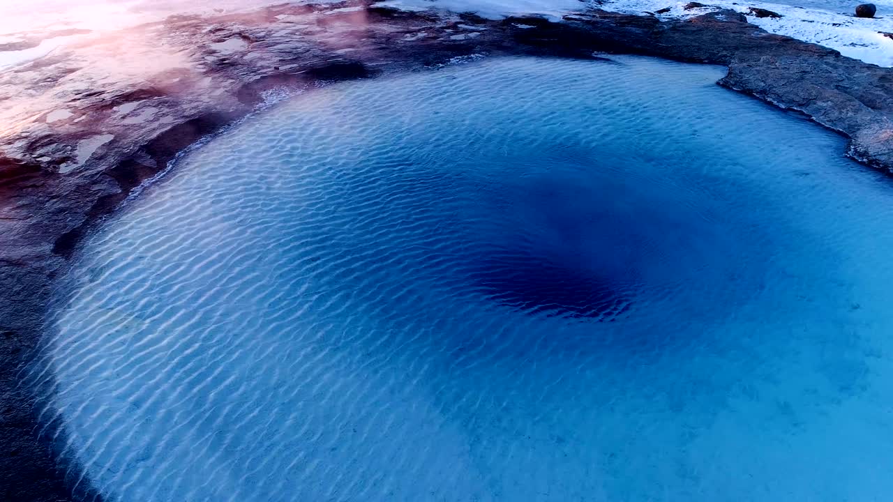 아이슬란드의 게이시르에 있는 게이저의 중심을 공중에서 볼 수 있습니다. 은 해가 지는 에 사람들이 게이저를 둘러싸고 고 있는 가운데 연기가 게이저 밖으로 나오고 있습니다.