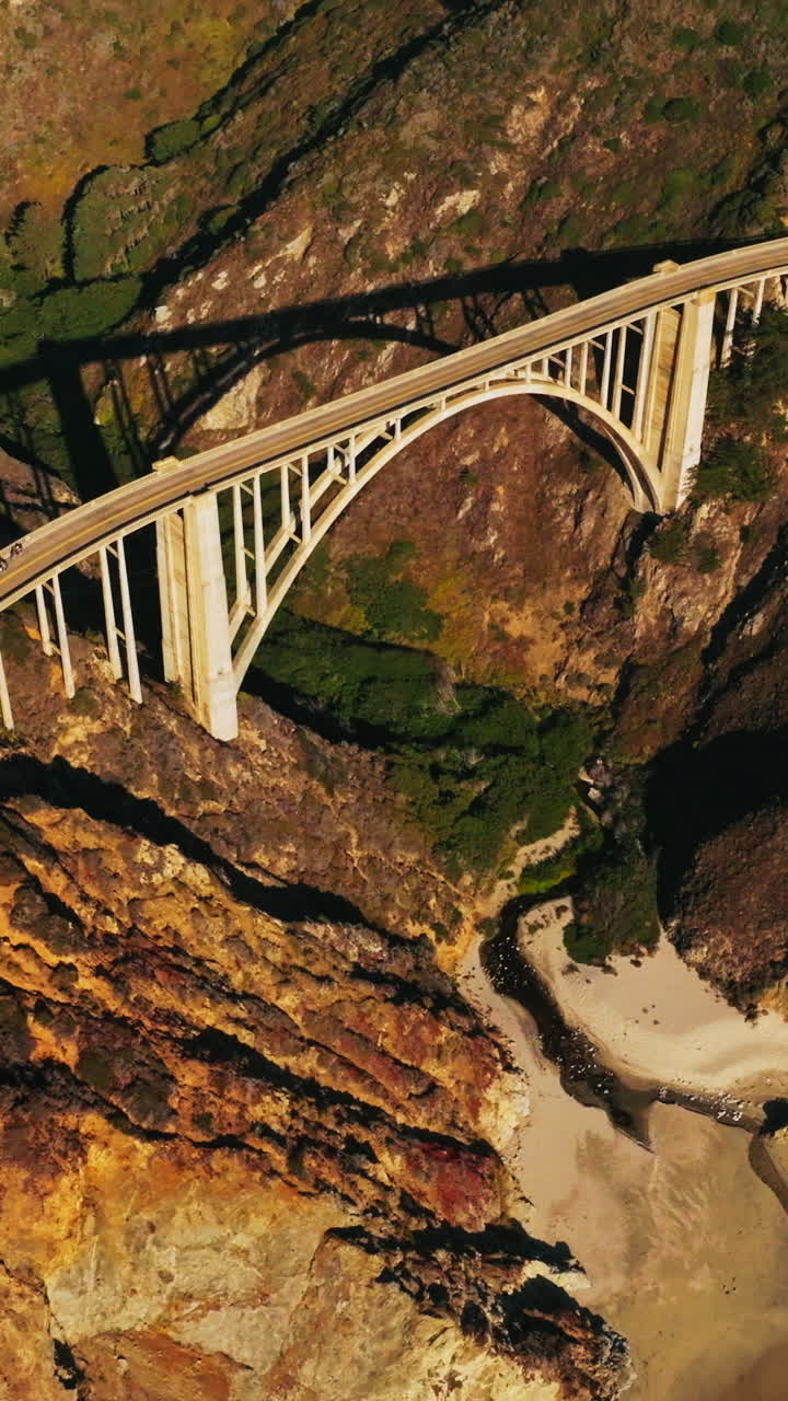 Sunlit rocks of the California coastline. Beautiful arched bridge connecting the mountains and highways on them. Aerial view. Vertical video