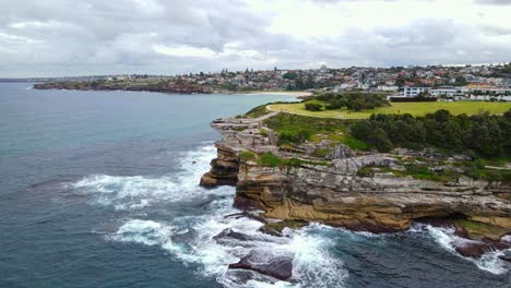 Olas-Rompiendo-En-El-Acantilado-Rocoso-De-Mackenzies-Point-Con-Turistas-En-El-Parque-De-Marcas-En-Tamarama,-Nsw,-Australia