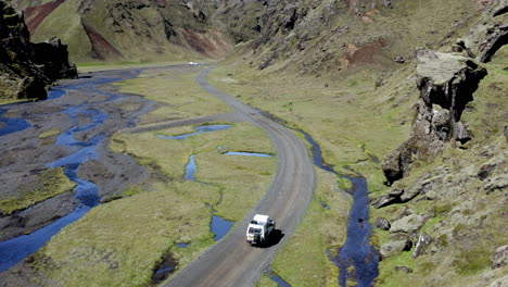 vista aérea de un camión blanco 4x4 conduciendo por el camino de tierra gris en el valle entre montañas rocosas y río ancho glaciar
