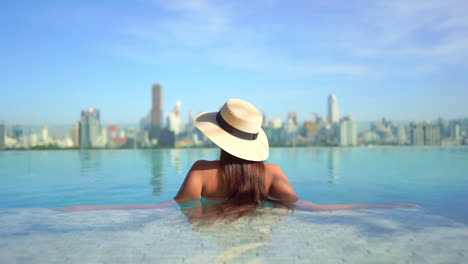 Backside-view-of-a-Pretty-young-model-wearing-a-swimming-suit-and-hat-and-sitting-inside-infinity-pool-enjoying-the-city-view,-hotels-rooftop