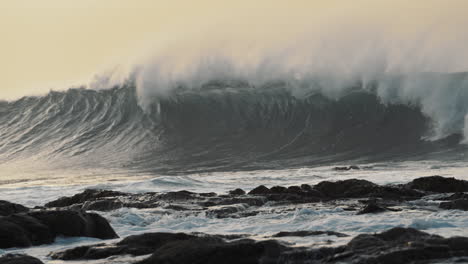 gran ola de surf en el océano rompiendo en cámara lenta con la costa rocosa en primer plano y el cielo naranja de la puesta de sol