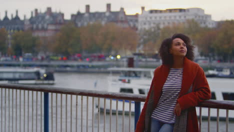 woman wearing winter coat leaning on railings by river thames in london at dusk - shot in slow motion