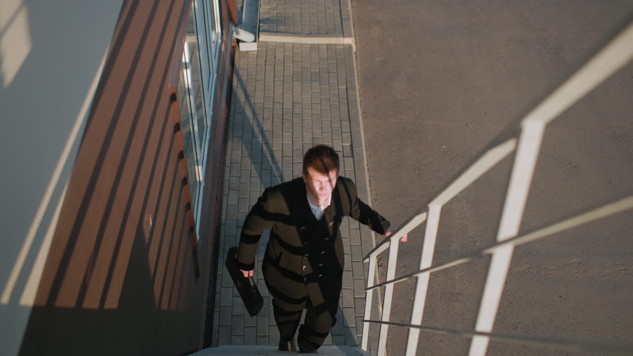 Man black suit climbing office staircase holding briefcase with right hand, iron railing with left hand, sunlight reflecting off face, focused, professional, upward movement