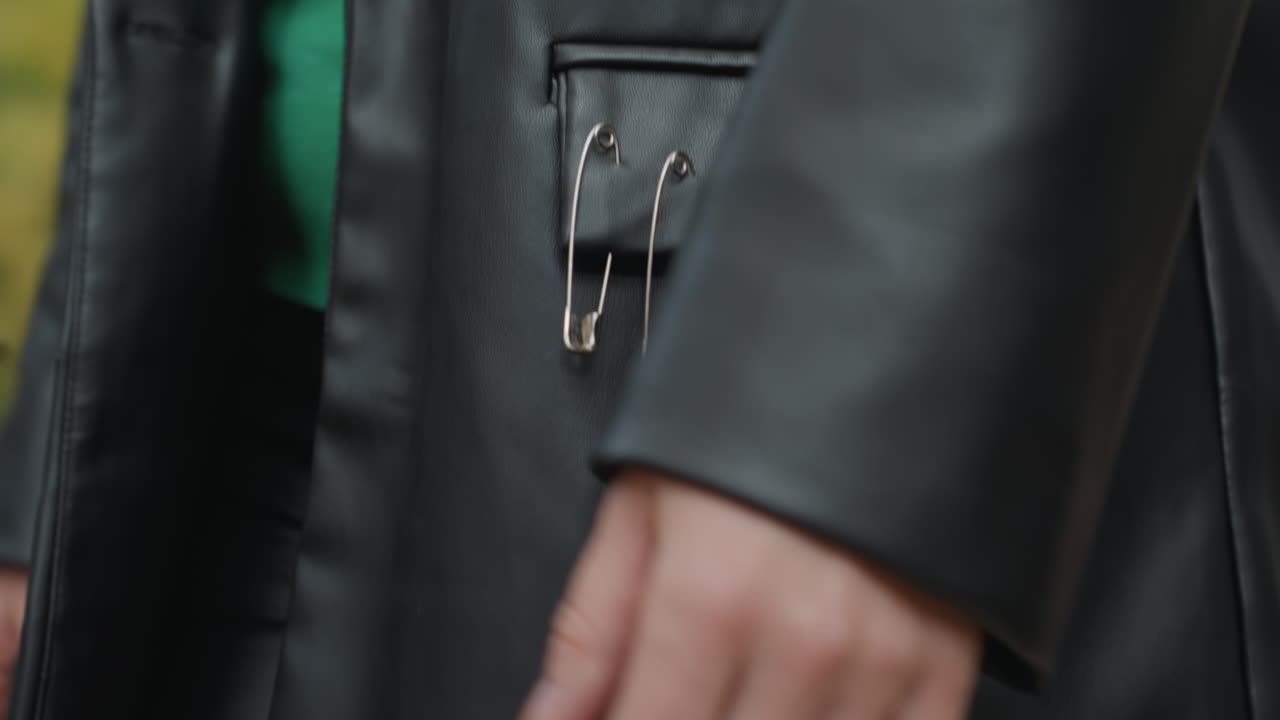 Nærbillede af læderjakkelomme med sikkerhedsnåle, kvinde står udendørs iført sort læderjakke med metalnåle fastgjort til lommen, grøn sweater titter frem i taljen, hånd afslappet ved siden, blød skov