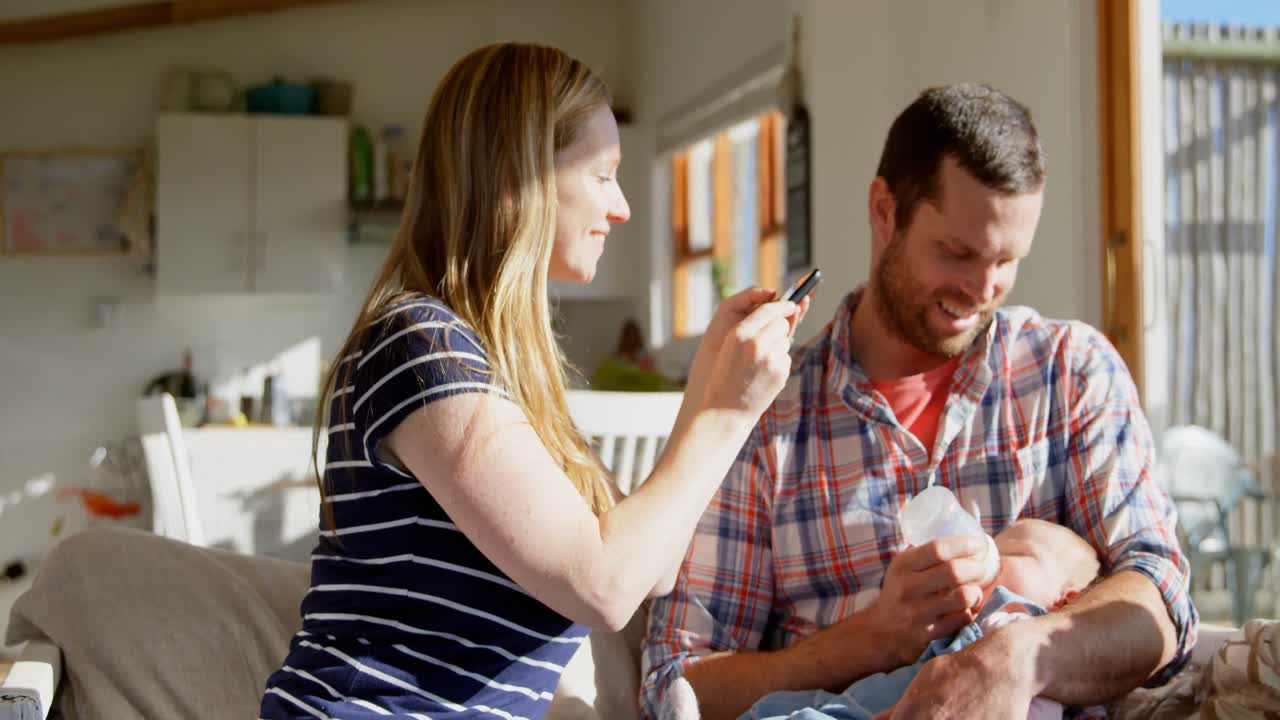 madre caucásica adulta haciendo clic en la foto mientras el padre alimenta a su bebé con leche del biberón en casa 4k