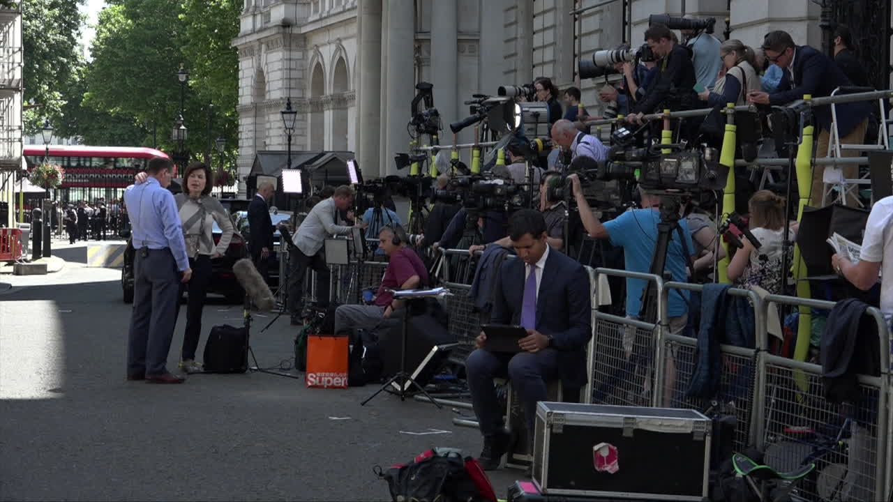 Two television news anchors talk to each as members of the press gather on Downing Street.