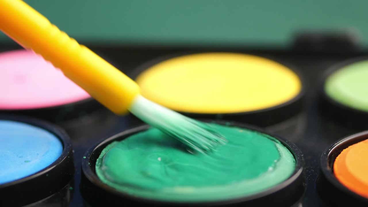 Paintbrush dipping into green watercolor paint in a colorful palette