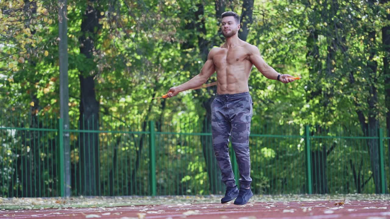 Fitness sportsman jumping with skipping rope. Muscular athlete doing exercise on rope on the outdoor stadium. Slow motion.