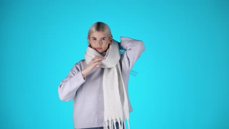 stylish young blonde woman, adjusting scarf and wearing grey sweater, studio shot with blue background