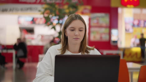 freelancer trabajando en una computadora portátil en el patio de comidas, escribiendo y concentrándose, sentado en una mesa en el interior de un centro comercial moderno con colores vibrantes, señalización de marca y actividad borrosa en el fondo