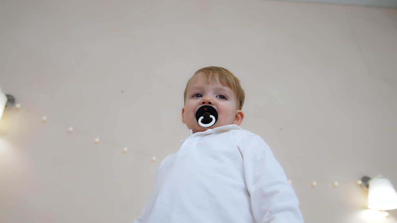 Beautiful toddler boy wearing white shirt and sucking a pacifier smiling adorably. Low angle view at the kid standing indoors.