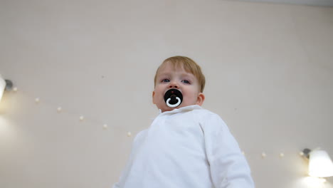 beautiful toddler boy wearing white shirt and sucking a pacifier smiling adorably. low angle view at the kid standing indoors.
