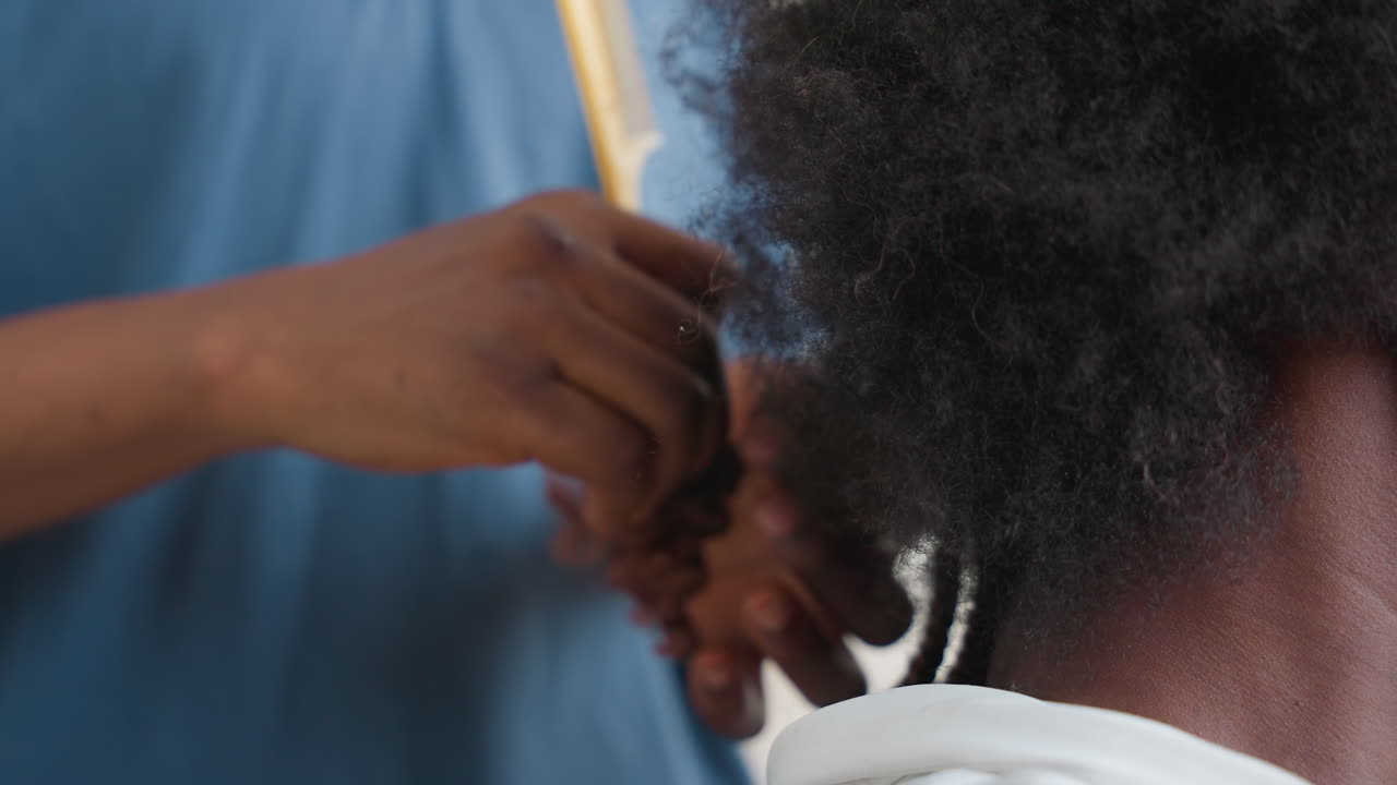 Primo piano di mani che intrecciano capelli neri in un ambiente domestico delicato. La stilista divide la nuca con pettine e dita, creando trecce strette e file texturizzate sotto una luce calda e soffusa. Cura tattile e tecnica paziente.