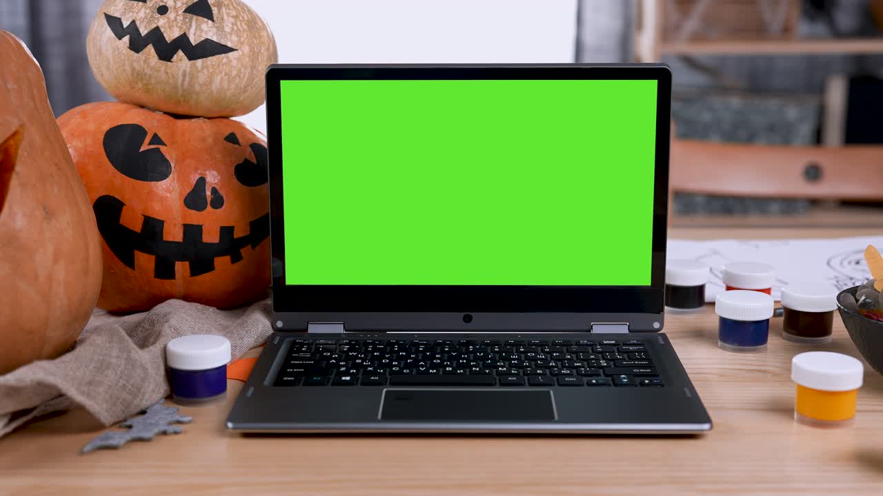 portátil moderno con maqueta de pantalla verde con llave de croma se encuentra en la mesa en la habitación. al lado del portátil hay calabazas cortadas con caras aterradoras, gouache y golosinas. zoom out. cámara lenta. cerrar