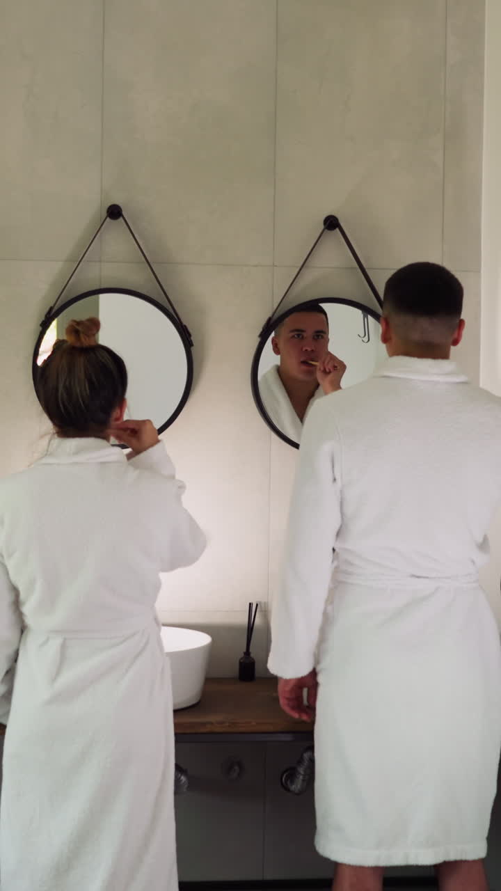 Lovers brush teeth in front of mirrors in bathroom. Couple in soft terry bathrobes performs everyday care procedures for dental health together