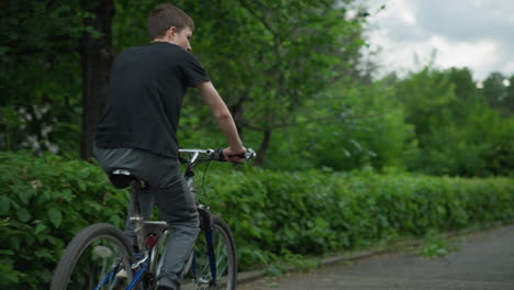 back view of two friends riding bicycles through lush green path, one standing while riding and the other sitting, the background features greenery with a blurred view of cars in the distance