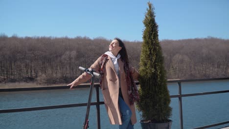 a young student girl in demi-season clothes with an electric scooter stands on an observation deck by the river and looks straight into the camera. front view. ecological city transport