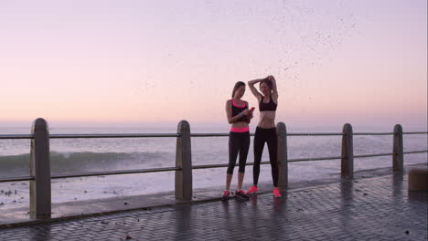 two athletic friends stretching before a run on promenade at sunset get wet from a wave