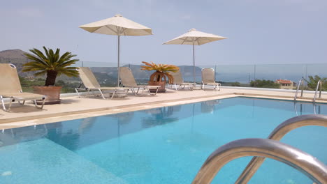 Gimbal-Shot-Pulling-Out-of-Sun-Loungers-on-Patio-of-Luxury-Greek-Villa-with-Pool-in-Foreground-and-Mountains-in-Background