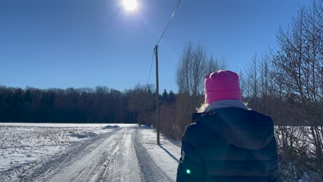 Frau-Geht-An-Einem-Sonnigen-Tag-Mit-Waldhintergrund-Im-Winter-Eine-Verschneite-Straße-Entlang