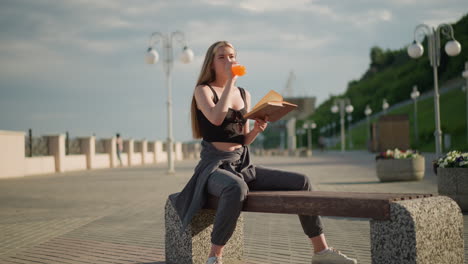 woman seated outdoors holding book in one hand while sipping juice from bottle with the other, focused on reading, the background features lamp posts and greenery