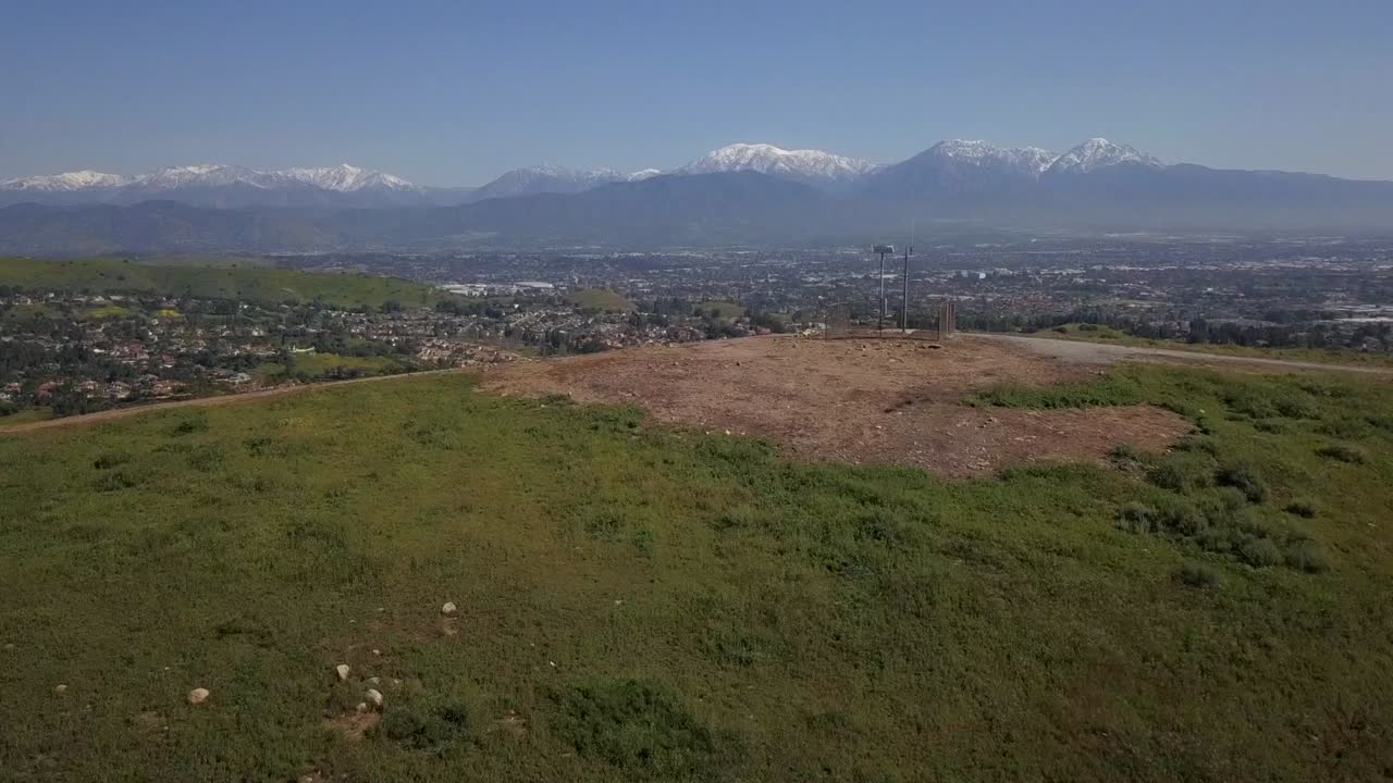 sobrevuelo aéreo de una colina cubierta de hierba que revela un amplio paisaje panorámico de la ciudad y autopistas con desarrollo urbano que alcanza las estribaciones del bosque nacional de angeles cubierto de nieve en la distancia
