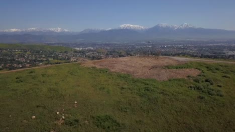 Aerial-flyover-of-a-grassy-hill-that-reveals-a-wide-panoramic-cityscape-and-freeways-with-urban-development-reaching-the-foothills-of-the-snowcapped-Angeles-National-Forest-in-the-distance