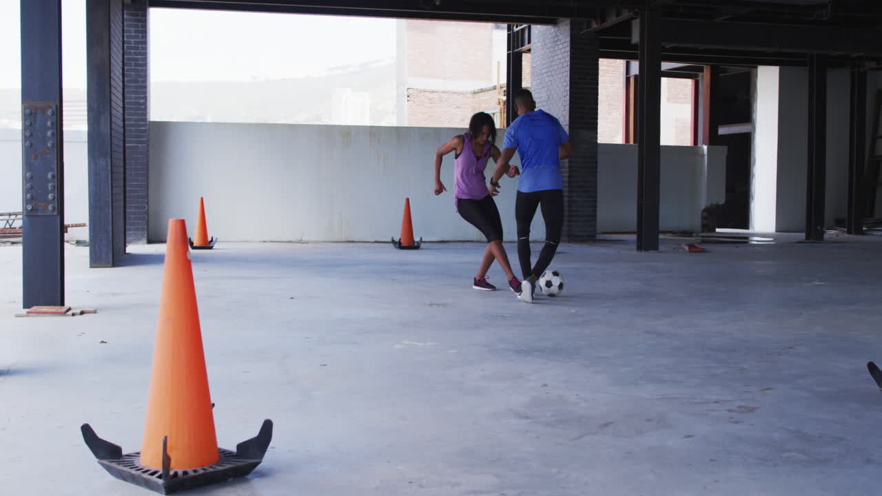 hombre y mujer afroamericanos jugando al fútbol en un edificio urbano vacío
