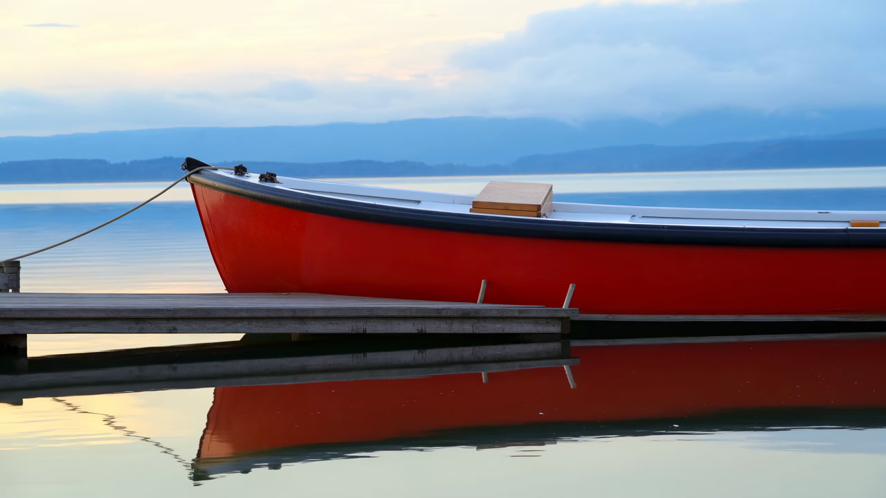 Red Boat Moored On Tranquil Lake At Dawn, Glassy Water Reflecting Hull And Misty Mountains. Wooden Dock