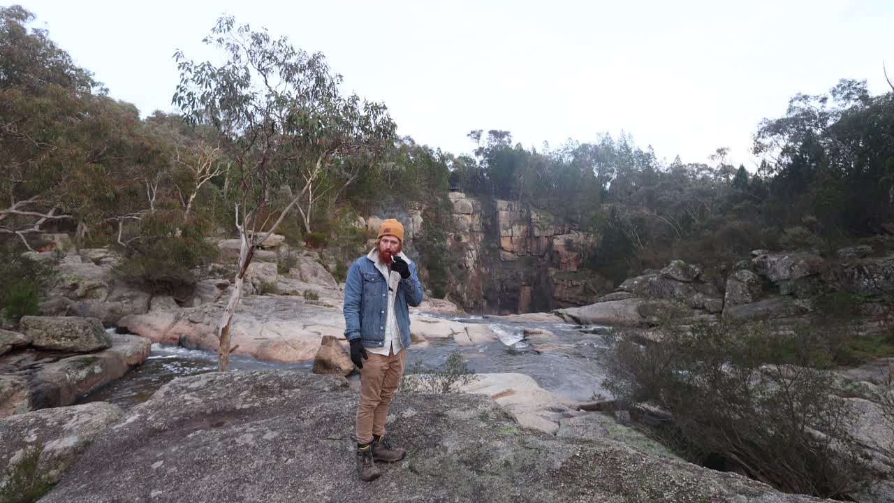 un bosquimano fumando una pipa y poniéndose guantes en el frío arbusto australiano encima de una cascada