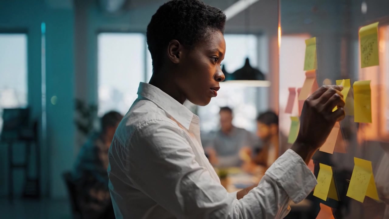 Focused woman engaging in creative brainstorming session, organizing colorful sticky notes on a transparent board in a modern office environment with teamwork present.