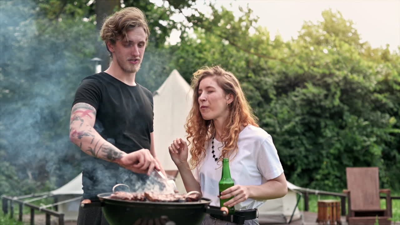 Giovane coppia che frigge carne sulla griglia, beve birra e la assaggia. Circondati dal verde. Glamping. Slow motion